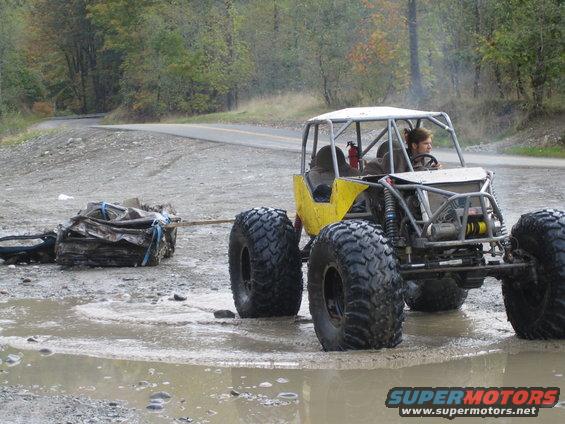 Yellow off-road buggy with roll cage and large tires tows a pile of scrap metal through a muddy puddle