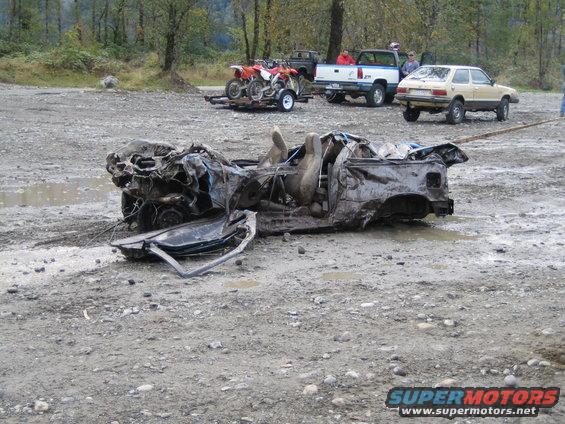 Wrecked car chassis with open doors and exposed interior, resting on a gravelly, muddy ground