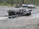 1990 Ford Bronco - A heavily damaged car chassis lies on the ground, with other vehicles and a trailer visible in the background.