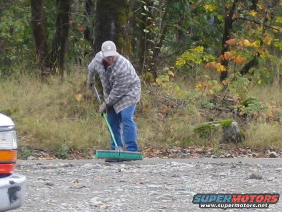 Man in plaid shirt and jeans sweeping a gravel area near trees