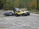 1990 Ford Bronco - Multiple off-road vehicles and a damaged pickup truck are parked on a gravel lot with trees in the background.