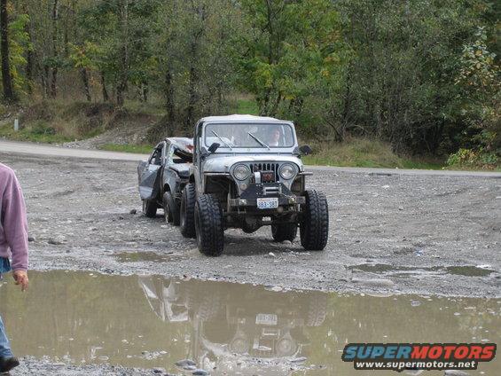 Jeep towing a beat-up car on a dirt road with trees in background. Puddle in foreground reflects the Jeep