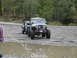 1990 Ford Bronco - A modified Jeep tows a damaged car through a muddy, gravelly area with trees lining the background.