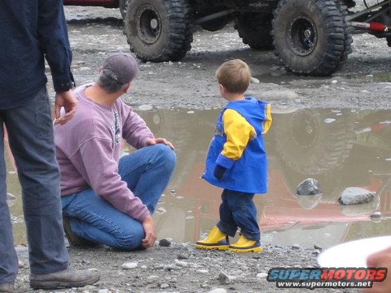 Man and child near a large tire in a muddy puddle; another off-road vehicle is partially visible