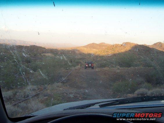 View from inside a vehicle looking at two off-road vehicles on a dirt path in a desert with hills