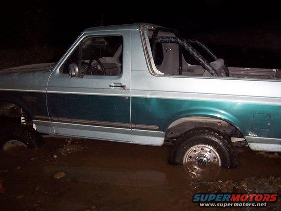 Side view of a 1996 Ford Bronco stuck in muddy water, with a roll cage visible inside