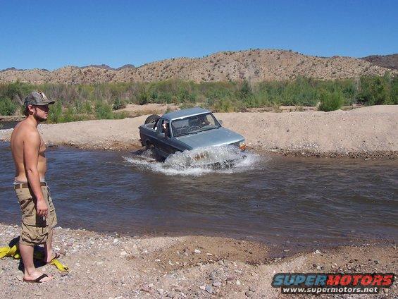 Light blue pickup truck with spare tire on back driving through shallow water, desert hills in background