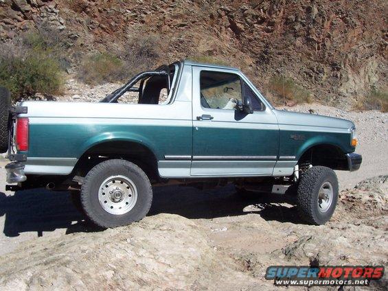 1996 Ford Bronco two-tone green and silver, lifted, off-road tires, spare tire on rear, driving on rocky terrain