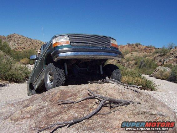 Green 1996 Ford Bronco climbing over a large rock on a dirt trail with desert hills in the background