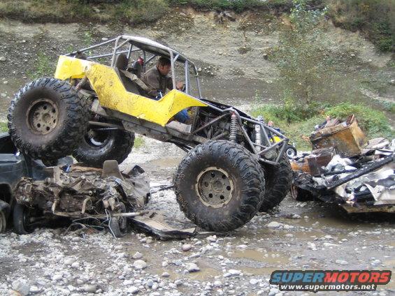 Yellow custom buggy with roll cage driving over wrecked cars in muddy terrain