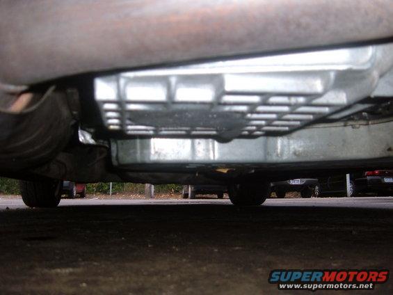 underside view of a car's transmission and exhaust system, with other vehicles visible in the background