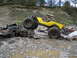 1990 Ford Bronco - A yellow off-road buggy is positioned precariously on top of a heavily damaged black pickup truck in a debris-filled, muddy area.
