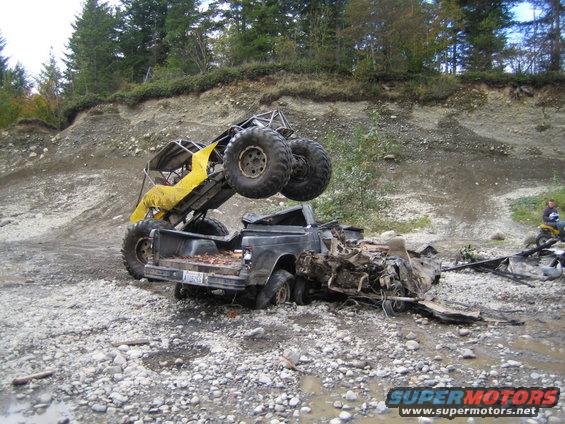 Yellow off-road buggy on top of a wrecked pickup truck in a muddy, rocky area