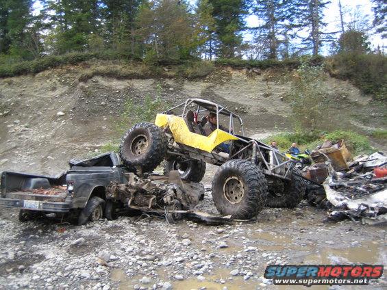 Yellow buggy on top of a wrecked black pickup truck in a muddy, rocky area. Another person is visible on a green ATV in the background