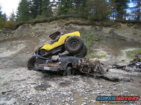 Yellow off-road vehicle on top of a wrecked black pickup truck, with a person inside the yellow vehicle
