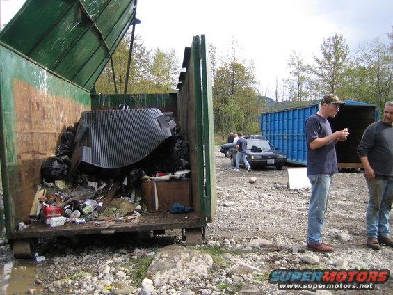 Dumpster filled with trash and a large black corrugated piece. Two men stand near a dark sedan and a blue shipping container