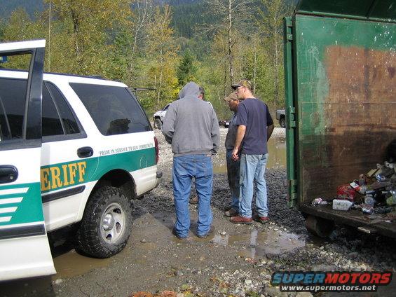 Sheriff SUV with open door, side view, parked in muddy area next to a dumpster