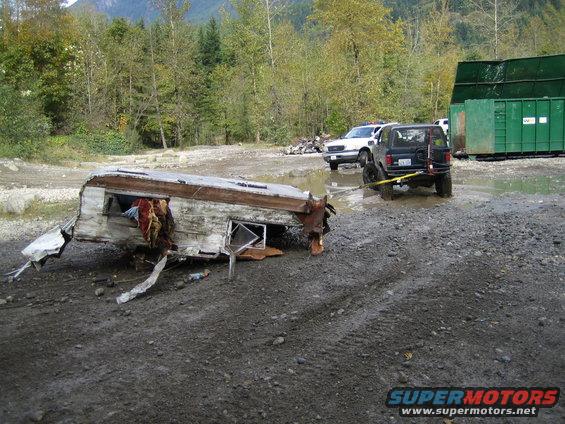 Black SUV towing damaged trailer, rear view. Supermotors.net logo visible