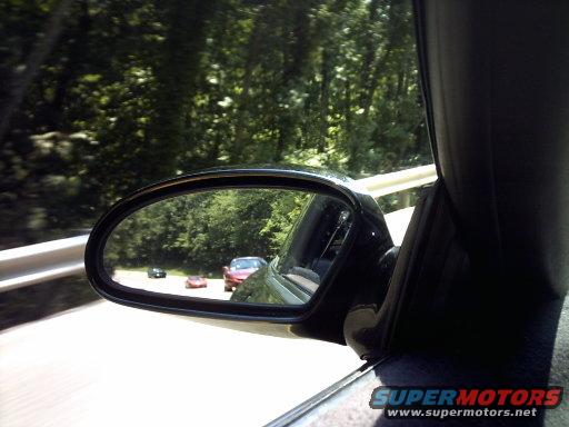 View of three cars on a road through a car's side mirror, with trees lining the road