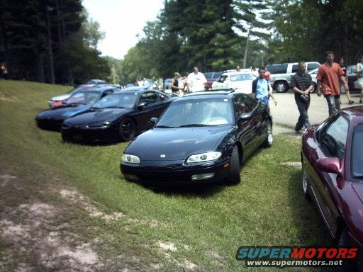 Several cars parked on a grassy area with trees in the background. A black coupe is in the foreground