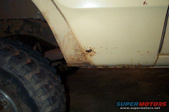 Close-up of a vehicle's side panel showing rust near the wheel well. A large tire is visible