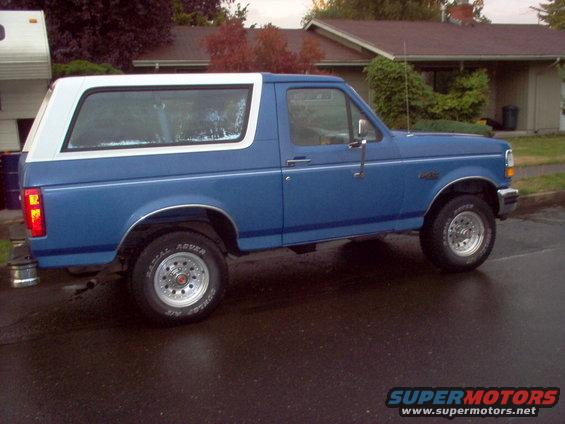 Blue 1992 Ford Bronco with a white top, seen from the side on a wet street