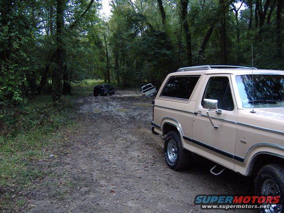 Tan 1984 Ford Bronco parked on a muddy dirt road; two other vehicles stuck in background