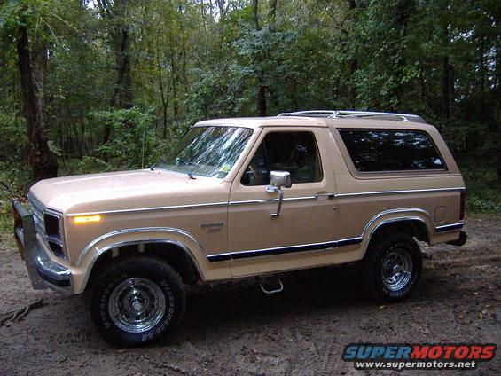 Tan 1984 Ford Bronco parked on dirt road with trees in background