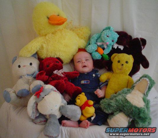 Baby in overalls sitting with a bunch of stuffed animals including a big yellow duck, teddy bears, and a green alligator