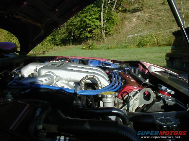 Engine bay with V6 intake manifold, blue spark plug wires, and a Supermotors.net watermark. Another car is visible in the background