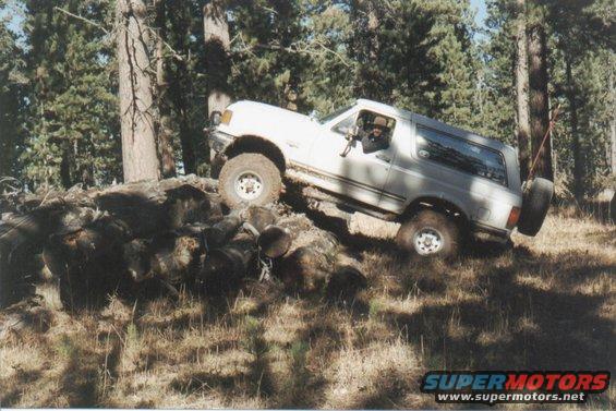 White Ford Bronco climbing over rocks and logs on a wooded trail