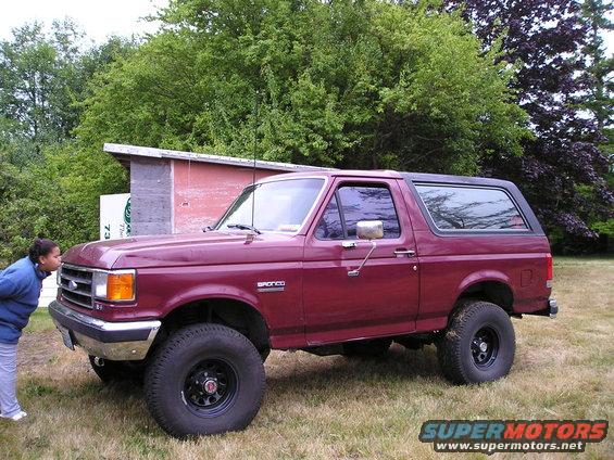 Maroon 1989 Ford Bronco parked on grass with a person leaning towards the front bumper