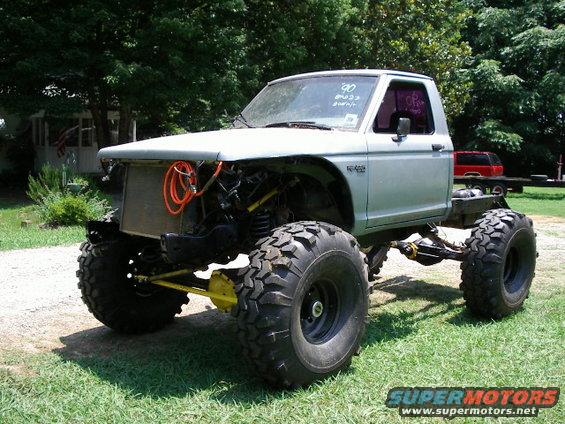 Modified pickup truck with large off-road tires, lifted suspension, and exposed engine components. A handwritten '90' is visible on the windshield