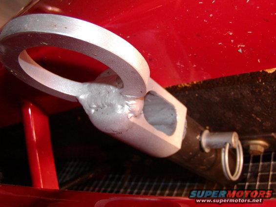 Close-up of a silver tow hook welded to a red car part, with a black tube and a ring attached