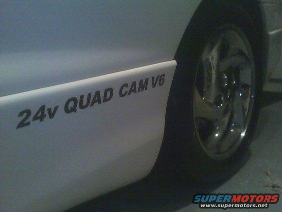 White car side with '24v QUAD CAM V6' decal and chrome wheel