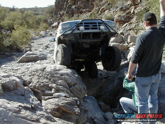 Ford Bronco with custom bumper driving over rocks. Two people are watching