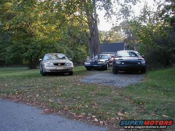 Three cars parked on a gravel driveway in front of a house. One is gold, one is black, and one is dark gray