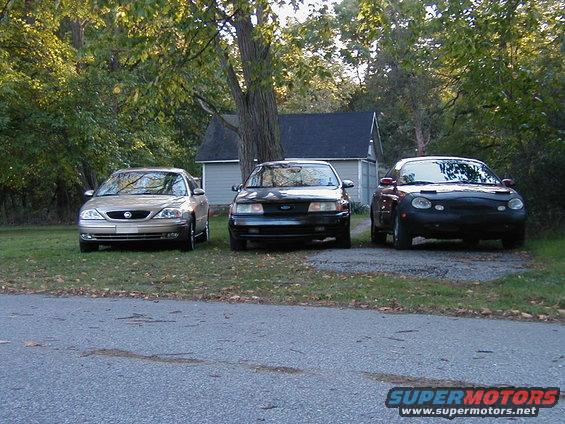 Three cars parked in front of a house, including a black 1990 Ford Taurus