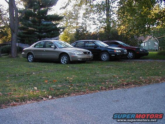 Three sedans parked on a grassy area next to a road, with trees and a house in the background