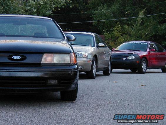Black Ford Taurus in front, silver sedan in middle, red car with cover in back