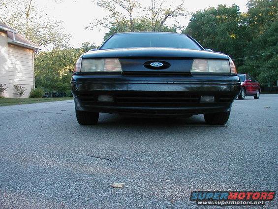 Front view of a black 1990 Ford Taurus parked on asphalt. Another red car is visible in the background