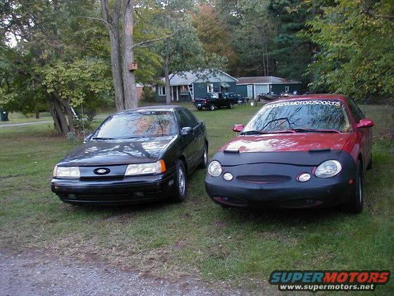 Black and red Ford Taurus sedans parked on grass. Red one has a car bra and a 'Florida Motorsports' banner on windshield