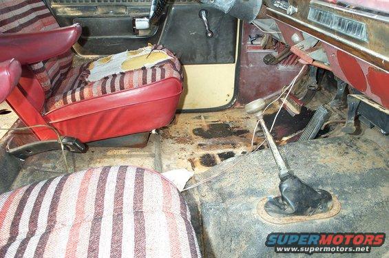 Interior of a vehicle with torn red and striped seats, rusty floor, and a floor shifter
