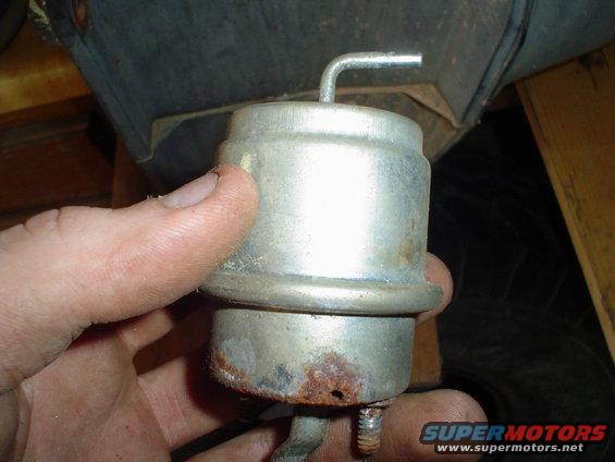 Hand holding a small, cylindrical metal vacuum actuator with a bent metal tube sticking out the top. Some rust is visible on the bottom