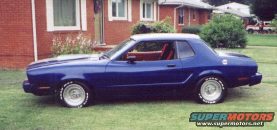 Blue coupe with red interior parked on grass in front of a brick house. Tires have white lettering