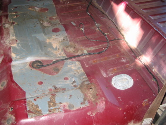 Rusted red floor pan with exposed metal patches and wiring. Visible fuel filler access