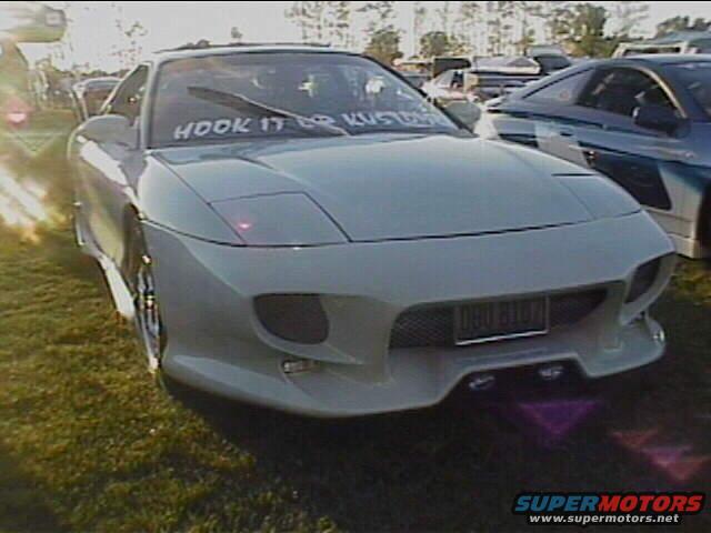Front view of a white sports coupe with a banner on the windshield that says 'HOOK IT UP KUSTOM'. License plate reads 'DBU 8107'. Another blue car is visible to the right