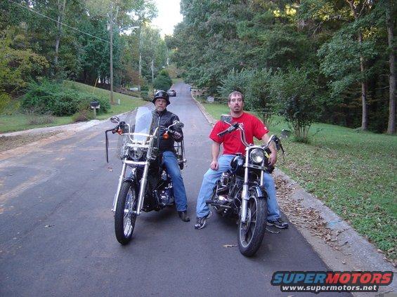 Two men sit on motorcycles on a tree-lined road. One man wears a black leather jacket, the other a red t-shirt. "SUPERMOTORS www.supermotors.net" is visible at the bottom right