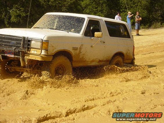 White 1987 Ford Bronco stuck in mud, front grille visible, mud splattered on windshield