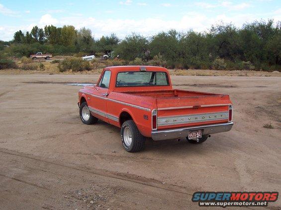 Orange pickup truck, rear 3/4 view, in a dirt lot. Chrome bumper, "CHEVROLET" tailgate